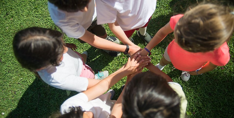 Flera barn står i en ring med utsträckta armar mot mitten en solig sommardag med grönt gräs.