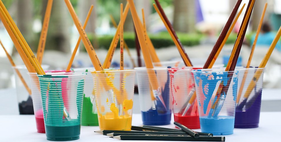 Flera plastglas står på ett bord med färgglada färger och penslar i varje glas.