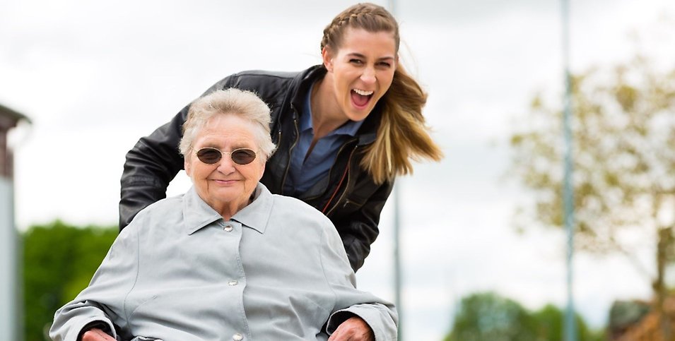 En äldre kvinna i rullstol tillsammans med en yngre kvinna, båda är glada.